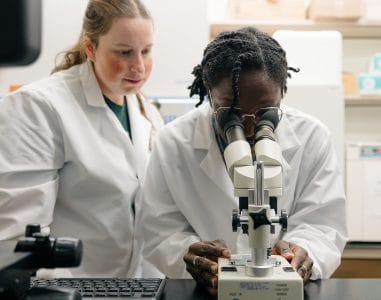 One scientist in a lab coat watching another scientist in a lab coat looking in a microscope